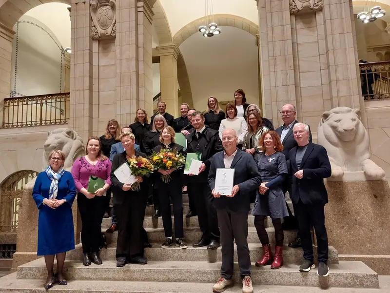 Festlich gekleidete Personen stehen auf den Stufen einer repräsentativen Säulenhalle, einige mit Blumensträußen oder Urkunden in den Händen