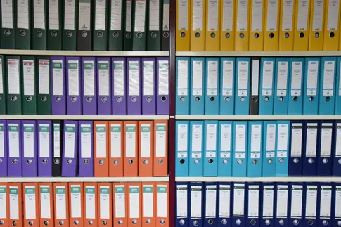 Viele verschiedenfarbige Aktenordner stehen, farblich sortiert, in einem Regal.