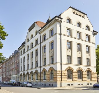 Sommerliche Aufnahme eines fünfstöckigen Gründerzeitgebäudes vor strahlend blauem Himmel. Das Gebäude ist ein Eckhaus und wird von zwei Straßen flankiert. Linker- und rechterhand stehen große grüne Laubbäume.