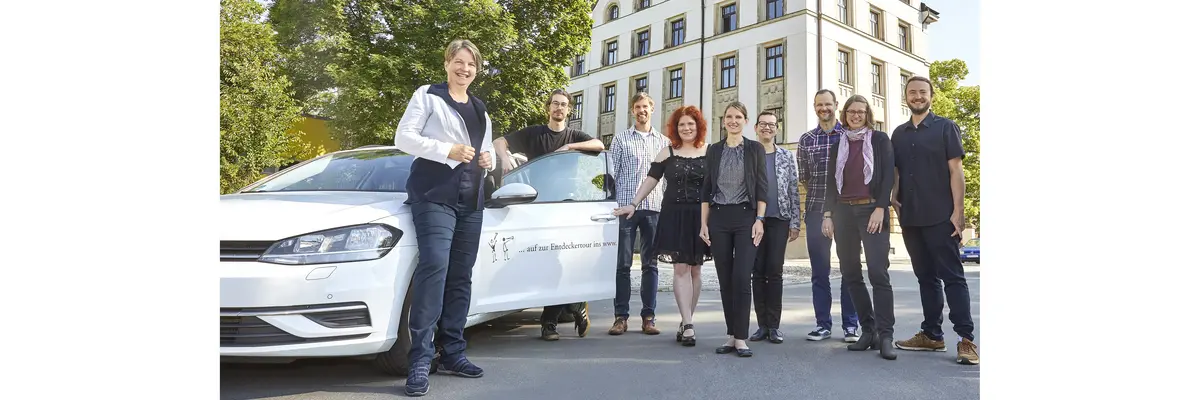 Neben einem weißen Auto mit der kleinen, seitlichen Aufschrift "auf zur Entdeckertour ins www" und geöffneter Fahrertür stehen fünf Frauen und vier Männer, die in die Kamera lächeln. Im Hintergrund ragen ein großer, grüner Laubbaum sowie ein fünfstöckiges Eckhaus aus der Gründerzeit auf.