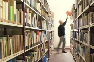 Eine Person steht in einem Gang einer Bibliothek. Links und rechts sind die Regale voll mit Büchern. Die Person nimmt sich ein Buch aus dem Regal.