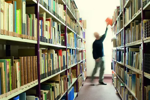 Eine Person steht in einem Gang einer Bibliothek. Links und rechts sind die Regale voll mit Büchern. Die Person nimmt sich ein Buch aus dem Regal.