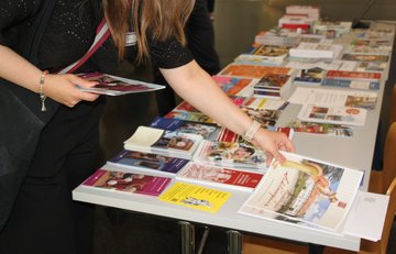 Nahaufnahme eines mit einer Vielzahl von Faltblättern bestückten Infotisches, an dem eine Person steht, von der nur Oberkörper, Arme und Hände zu sehen sind. Die Person hat bereits ein paar Flyer in einer Hand und greift mit der anderen Hand eine weitere Broschüre.