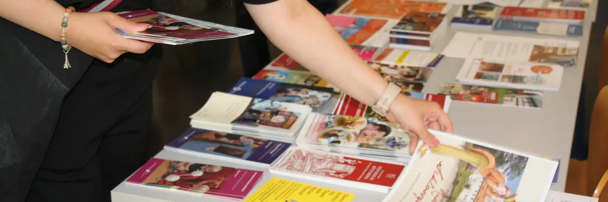 Nahaufnahme eines mit einer Vielzahl von Faltblättern bestückten Infotisches, an dem eine Person steht, von der nur Oberkörper, Arme und Hände zu sehen sind. Die Person hat bereits ein paar Flyer in einer Hand und greift mit der anderen Hand eine weitere Broschüre.