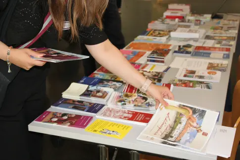 Nahaufnahme eines mit einer Vielzahl von Faltblättern bestückten Infotisches, an dem eine Person steht, von der nur Oberkörper, Arme und Hände zu sehen sind. Die Person hat bereits ein paar Flyer in einer Hand und greift mit der anderen Hand eine weitere Broschüre.