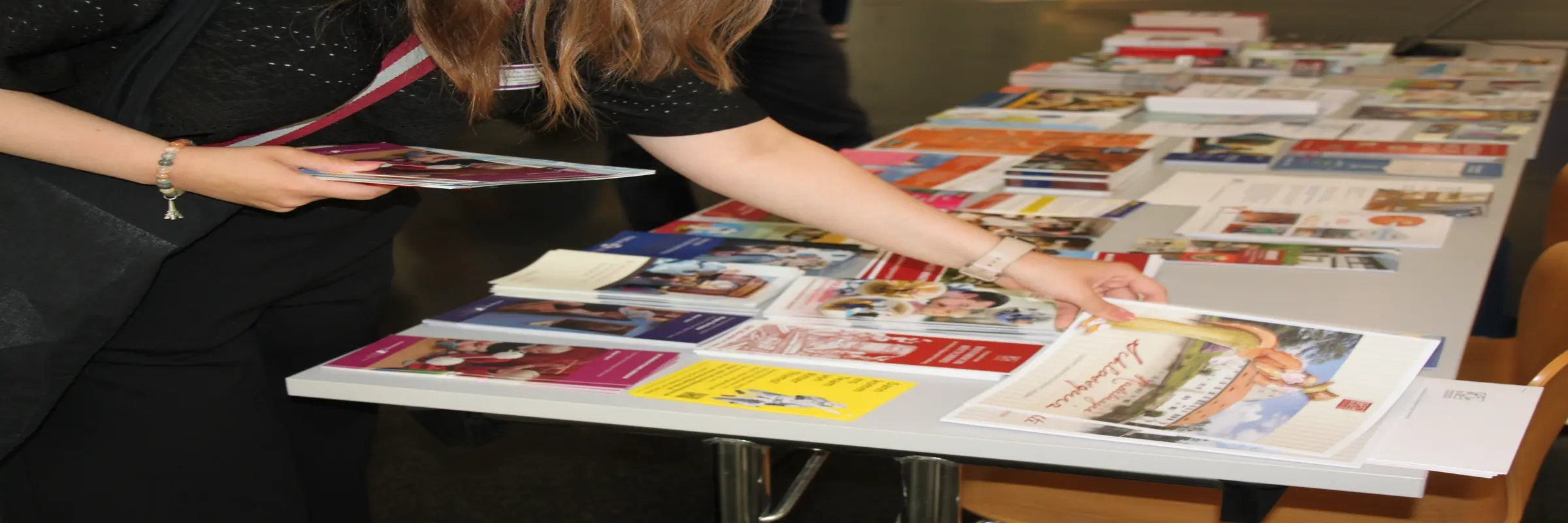Nahaufnahme eines mit einer Vielzahl von Faltblättern bestückten Infotisches, an dem eine Person steht, von der nur Oberkörper, Arme und Hände zu sehen sind. Die Person hat bereits ein paar Flyer in einer Hand und greift mit der anderen Hand eine weitere Broschüre.