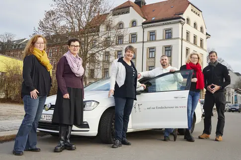 Neben einem weißen Auto mit der seitlichen Aufschrift "Auf zur Entdeckertour ins www" und geöffneter Fahrertür stehen vier Frauen und zwei Männer; im Hintergrund ein fünfstöckiges Gründerzeit-Eckhaus