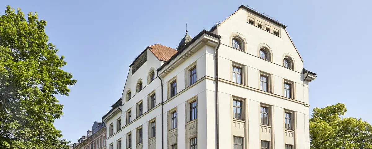 Sommerliche Aufnahme eines fünfstöckigen Gründerzeitgebäudes vor strahlend blauem Himmel. Das Gebäude ist ein Eckhaus und wird von zwei Straßen flankiert. Linker- und rechterhand stehen große grüne Laubbäume.