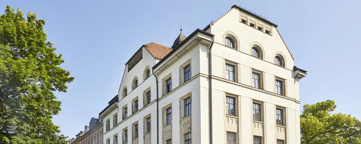 Sommerliche Aufnahme eines fünfstöckigen Gründerzeitgebäudes vor strahlend blauem Himmel. Das Gebäude ist ein Eckhaus und wird von zwei Straßen flankiert. Linker- und rechterhand stehen große grüne Laubbäume.